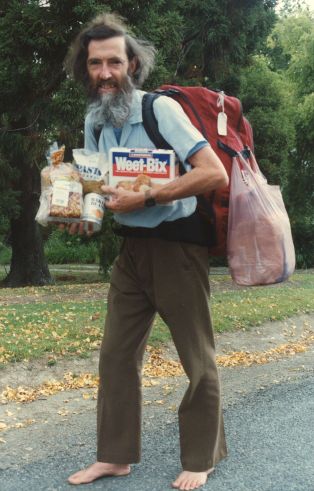 [colour photo, David MacClement on road: long dark hair, long beard, red backpack, bare feet]