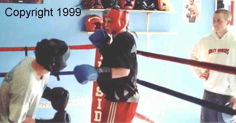 Outside the ring, a young boxer watches the sparring.  He is still learning the basics and has yet to enter the ring, but will soon!  He can learn much by watching the match and listening to the the critique of the coach.  Image copyright 1999.  All rights reserved.