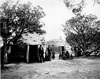 13 NAVAJO INDIVIDUALS STANDING BY FRED HARVEY NAVAJO EMPLOYEE HOGANS ADJOINING HOPI HOUSE & EL TOVAR HOTEL. CIRCA 1906. FRED HARVEY CO. LOC # H-80610