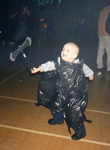 Excited smiling boy with no hair dressed in bin bag and waving toy axe