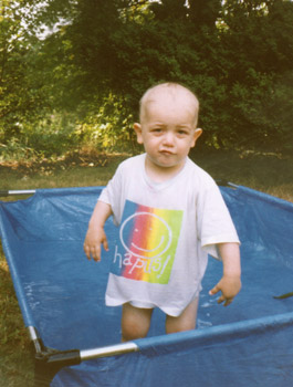 little boy with no hair standing in paddling pool wearing rainbow T-shirt with Welsh word happis for happy. His expression is a mock scowl